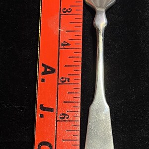 May include: A silver-toned metal fork with four tines and a smooth, flared handle, positioned next to a bright orange ruler. The fork measures approximately 7.5 inches in length, as indicated by the markings on the ruler placed alongside it.