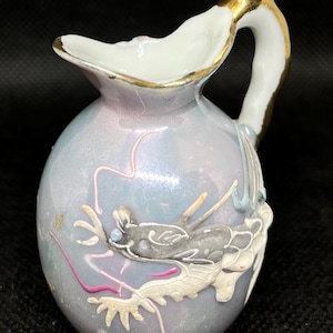 May include: A small porcelain pitcher with an iridescent light blue finish, featuring a raised relief of a grey dragon with pink accents. The handle and the rim of the spout are accented with gold trim, and the body has a smooth, glossy texture.