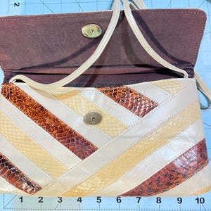 May include: A rectangular shoulder bag featuring a diagonal patchwork design of cream, tan, and brown snakeskin-textured leather. The bag has a gold-tone metal clasp, a thin shoulder strap, and a dark brown interior lining, measuring 10 inches wide.