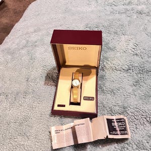 May include: A gold-tone Seiko wristwatch with a white dial, presented in a maroon and cream-colored box with the Seiko logo. A folded paper instruction manual and warranty certificate are placed in front of the open box on a textured surface.