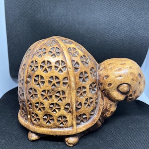 May include: A brown turtle figurine with a textured shell featuring a repeating geometric star pattern. The head and limbs have a smooth, rounded appearance with subtle dark brown shading in the crevices of the carved shell design.