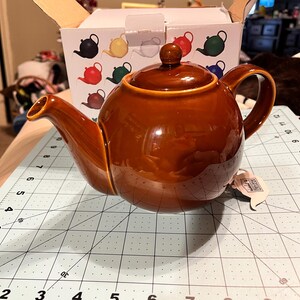 May include: A glossy brown ceramic teapot with a matching lid, positioned in front of its original cardboard box. The box features illustrations of teapots in various colors. A white tag is attached to the handle of the teapot.