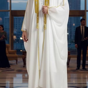 Könnte beinhalten: Ein weißer Bisht mit goldener Stickerei entlang der vorderen Öffnung und des Kragens. Das Kleidungsstück ist ein langer, fließender, vorne offener Umhang mit weiten Ärmeln, der über einem weißen Thobe getragen wird. Das Wort WHITE steht oben links.