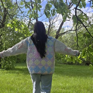 May include: A pastel-coloured quilted jacket with a diamond pattern. The jacket is light blue, pink, and yellow. The person is wearing light blue jeans and standing in a grassy area with trees.