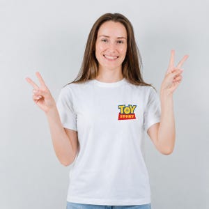 Może przedstawiać: Biała koszulka z logo "Toy Story" w kolorze ż&oacute;łtym, niebieskim i czerwonym. Koszulka ma kr&oacute;tkie rękawy i jest wykonana z miękkiego materiału. Logo znajduje się na lewej piersi.