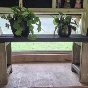 May include: A small, dark gray wooden bench with light brown legs. Two potted plants sit on the bench in front of a window. The bench is made of wood and has a simple, rustic design, suitable for indoor or outdoor use.