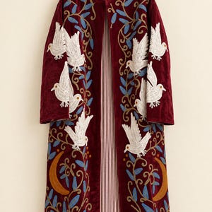 May include: A long, deep red velvet coat featuring intricate embroidery of white birds, blue leaves, and golden crescent moons. The open-front garment has a mandarin collar and is lined with a white and red vertical pinstriped fabric.
