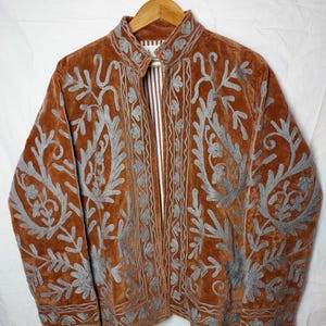 Puede incluir: Una chaqueta de color óxido con intrincados bordados de color gris plateado en un patrón floral y de cachemira. La chaqueta tiene cuello alto, mangas largas y una percha de madera marrón. El forro interior es visible y presenta rayas blancas y marrones.