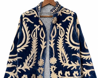 May include: A deep blue velvet jacket featuring intricate cream-colored embroidery in a swirling paisley and floral pattern. The garment has long sleeves, a high collar, and an open front that reveals a black and white vertical striped lining.