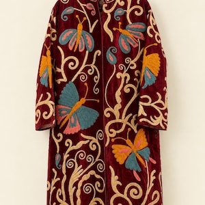 May include: A deep burgundy velvet coat featuring intricate embroidery of butterflies and swirling vine patterns in shades of teal, orange, and cream. The long-sleeved garment has a high collar and a straight, open-front silhouette.