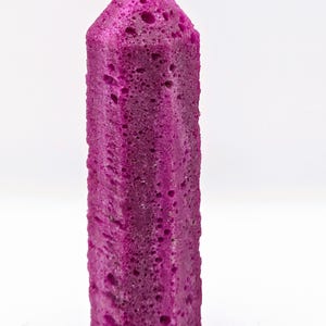 May include: A vibrant magenta, hexagonal crystal point with a porous, pitted surface texture. The object features a pointed top and a flat base, displaying a consistent deep pink color throughout its elongated, vertical, and faceted structure.