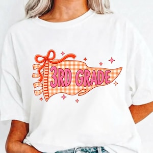 Könnte beinhalten: Ein weißes T-Shirt mit einer Grafik eines orange-weiß karierten Wimpels mit einer roten Schleife. Der Text 3RD GRADE ist in Pink mit roter Umrandung im Wimpel gedruckt und von kleinen roten Sternen auf dem Vorderteil des Designs umgeben.