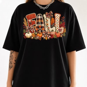 Puede incluir: Una camiseta negra con un gráfico grande en el pecho que forma la palabra FALL. Las letras están decoradas con diversas texturas, incluyendo patrones de punto, cuadros y motivos florales, rodeadas de calabazas, lazos y hojas de otoño.