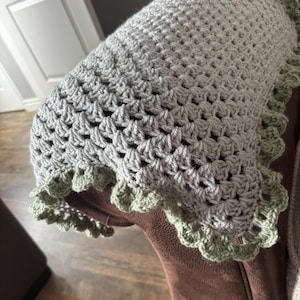 May include: A handmade crochet blanket in shades of grey with a scalloped green border. The blanket features a textured stitch pattern, creating a cosy and inviting appearance. The soft colour palette and intricate design suggest a focus on comfort and craftsmanship.