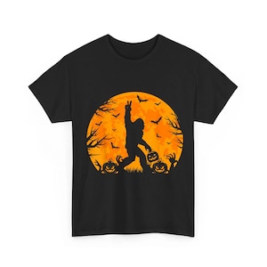 May include: A black short-sleeve t-shirt featuring a graphic of a silhouette of Bigfoot making a peace sign against a large orange full moon. The design includes flying bats, bare tree branches, and two jack-o'-lanterns on the ground.