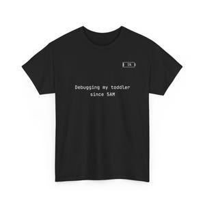 May include: A black short-sleeved t-shirt featuring a white graphic of a battery icon at 1% charge and the white text "Debugging my toddler since 5AM" printed in a clean, sans-serif font across the center of the chest area.
