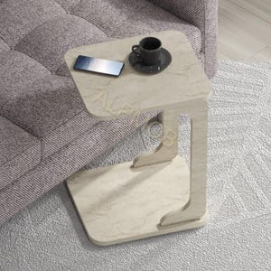 Könnte beinhalten: Ein beigefarbener Beistelltisch in Marmoroptik mit C-förmigem Design, auf dem eine schwarze Kaffeetasse und ein Smartphone stehen. Der Tisch steht neben einem grauen Sofa auf einem hellgrauen Teppich.