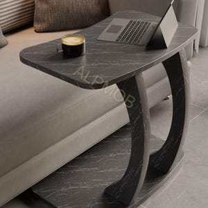 May include: A C-shaped side table with a black faux marble finish and white veining. The table features a rectangular top, a curved support base, and a flat bottom platform. The text ALP MOB is printed on the surface of the table top.