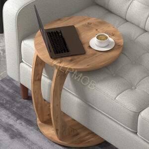 Könnte beinhalten: Ein runder Beistelltisch aus Holz mit einem Laptop und einer Kaffeetasse. Der Tisch hat eine hellbraune Oberfläche und einen einzigartigen geschwungenen Fuß. Er steht neben einem grauen Sofa und schafft so einen gemütlichen und funktionalen Raum.