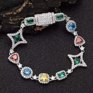 May include: A silver-toned link bracelet featuring a central rectangular clasp encrusted with small clear stones. The chain alternates between round, triangular, and star-shaped settings containing multi-colored gemstones in red, blue, green, and yellow.
