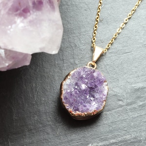 A purple Amethyst Geode Cluster necklace lays flat on a piece of grey slate. The points of the raw crystal sparkle in the sunlight. A band of gold plating covers the edge of the crystal. To the left of the pendant is a large piece of Amethyst.