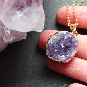 A purple Amethyst Geode Cluster necklace lays flat on a hand, covering 2 fingers. The points of the raw crystal sparkle in the sunlight. A band of gold plating covers the edge of the crystal. To the left of the pendant is a large piece of Amethyst.