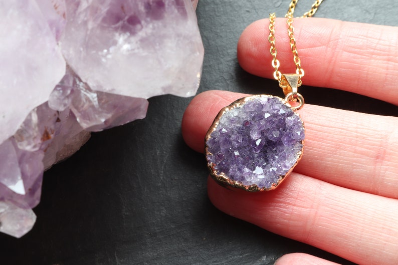 A purple Amethyst Geode Cluster necklace lays flat on a hand, covering 2 fingers. The points of the raw crystal sparkle in the sunlight. A band of gold plating covers the edge of the crystal. To the left of the pendant is a large piece of Amethyst.