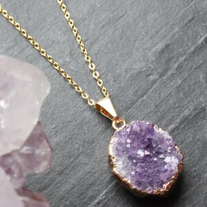 A purple Amethyst Geode Cluster necklace lays flat on a piece of grey slate. The points of the raw crystal sparkle in the sunlight. A band of gold plating covers the edge of the crystal. To the left of the pendant is a large piece of Amethyst.