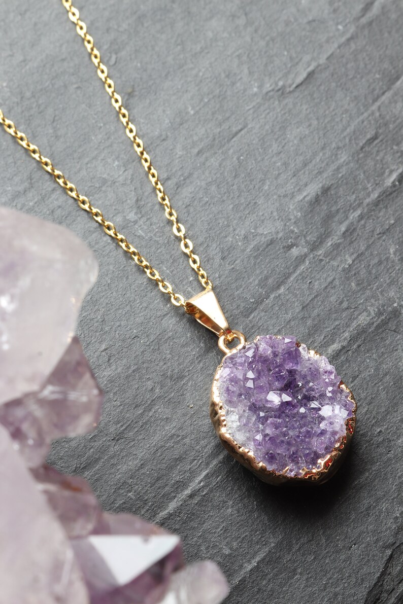 A purple Amethyst Geode Cluster necklace lays flat on a piece of grey slate. The points of the raw crystal sparkle in the sunlight. A band of gold plating covers the edge of the crystal. To the left of the pendant is a large piece of Amethyst.