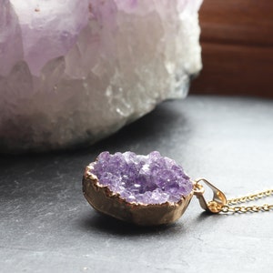 A side on view of an Amethyst Geode Cluster necklace laying flat on a piece of grey slate. The points of the raw crystal sparkle in the sunlight. A band of gold plating covers the edge of the crystal.