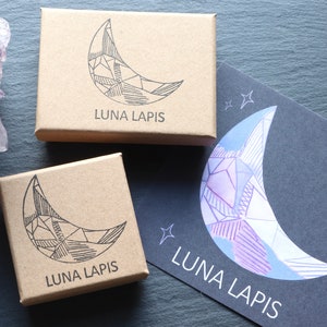 2 brown card jewellery boxes, hand stamped with the LUNA LAPIS logo, alongside a LUNA LAPIS branded postcard. The logo depicts a crystal in the shape of a crescent moon. To the left of the boxes is a large Amethyst Cluster crystal.