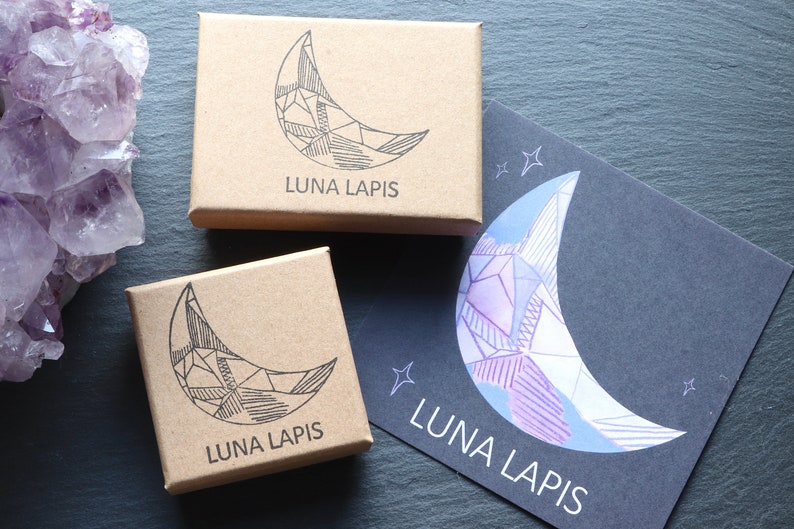 2 brown card jewellery boxes, hand stamped with the LUNA LAPIS logo, alongside a LUNA LAPIS branded postcard. The logo depicts a crystal in the shape of a crescent moon. To the left of the boxes is a large Amethyst Cluster crystal.