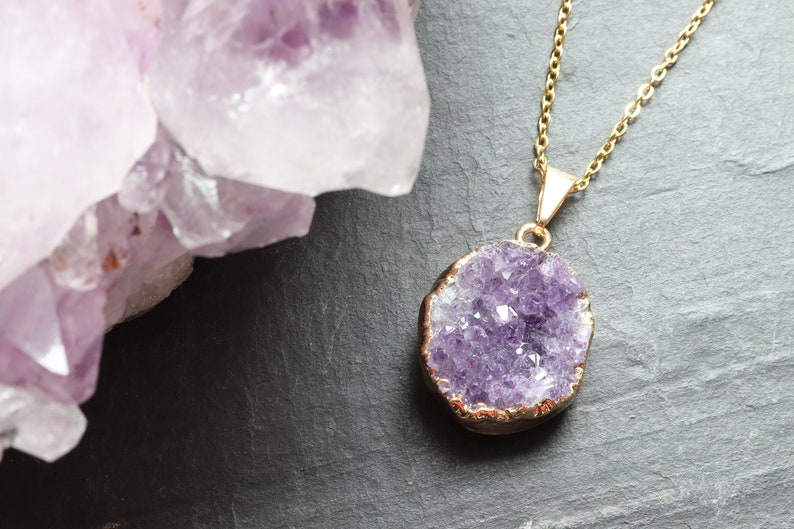 A purple Amethyst Geode Cluster necklace lays flat on a piece of grey slate. The points of the raw crystal sparkle in the sunlight. A band of gold plating covers the edge of the crystal. To the left of the pendant is a large piece of Amethyst.