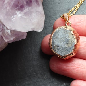 Image shows the back of an Amethyst Geode Cluster necklace, held in a hand. The smooth stone is exposed on the back of the pendant. A band of gold plating covers the edge of the crystal. To the left of the pendant is a large piece of Amethyst.