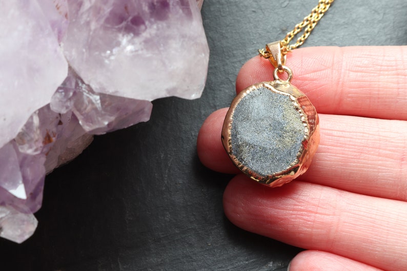 Image shows the back of an Amethyst Geode Cluster necklace, held in a hand. The smooth stone is exposed on the back of the pendant. A band of gold plating covers the edge of the crystal. To the left of the pendant is a large piece of Amethyst.