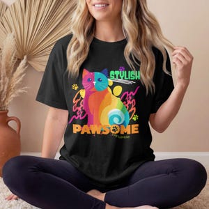 Puede incluir: Una camiseta negra de manga corta con un gráfico de gato multicolor y vibrante. El diseño incluye el texto STYLISH, PAWSOME y CAT LOVERS en letras negritas, acentuado con pequeñas huellas de patas y formas abstractas en amarillo, rosa y azul.