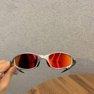 May include: Silver-toned metal sunglasses featuring a wrap-around frame design and reflective red lenses. The eyewear has a distinct metallic bridge and contoured temples, showcasing a sleek, aerodynamic construction with vibrant crimson-tinted glass.