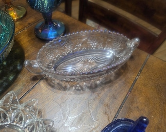 Puede incluir: Un plato ovalado de cristal transparente con dos pequeñas asas en los laterales. El plato presenta un patrón en relieve de uvas y hojas en el borde y un diseño de rayos de sol en la base interior. Los bordes son festoneados y con cuentas.