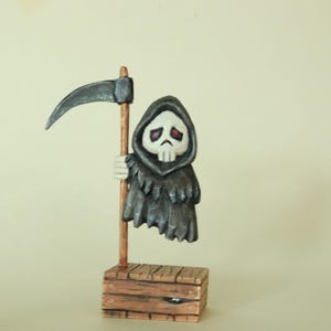 May include: A small figurine of a grim reaper character with a white skull face, red eyes, and a dark grey hooded cloak. The figure holds a long wooden scythe and stands on a rectangular wooden crate base with visible grain and dark brown accents.