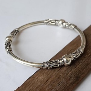 May include: A silver bangle bracelet featuring a smooth, circular band accented with three polished spherical beads and three elongated, textured metal segments with intricate, openwork detailing arranged symmetrically around the circumference.