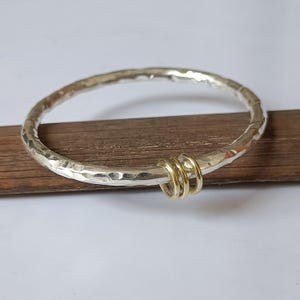 Puede incluir: Un brazalete de color plateado con una superficie martillada y texturizada. El brazalete presenta tres anillos pequeños, lisos y de color dorado que están enlazados alrededor de la banda principal, permitiéndoles deslizarse libremente.