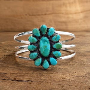 May include: A silver cuff bracelet featuring a large central oval turquoise stone surrounded by twelve smaller teardrop-shaped turquoise stones. The metal band consists of three thin, polished silver wires that meet at the sides to support the cluster.
