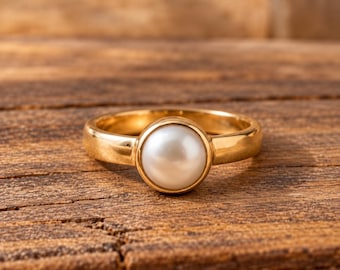 Könnte beinhalten: Ein Goldring mit einer einzelnen, runden, weißen Perle, die in eine glatte, polierte Goldschiene gefasst ist. Die Perle ist in einer Zargenfassung zentriert, was ein klares, kreisförmiges Profil erzeugt. Das Metall hat ein warmes Finish.
