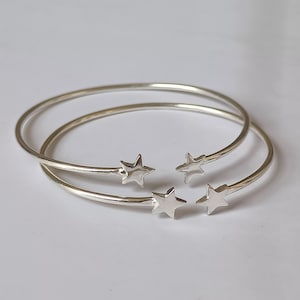 May include: Two silver-toned open cuff bracelets, each featuring a polished star motif at both ends. The bracelets are circular in shape with a smooth, reflective metallic finish, designed as open-ended bangles that showcase a star on each terminal.