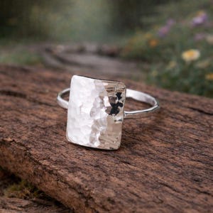 May include: A silver ring featuring a rectangular face with a hammered, textured surface. The band is thin and polished, connecting to the sides of the rectangular centerpiece. The metal has a bright, reflective finish that catches the light.