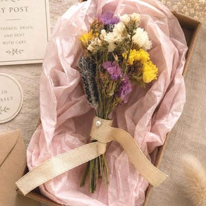 Peut inclure: Un petit bouquet de fleurs séchées composé de fleurs jaunes, blanches et violettes, noué par un ruban beige agrémenté d'une perle. Le bouquet est placé dans une boîte en carton brun tapissée de papier de soie rose pâle et délicat.