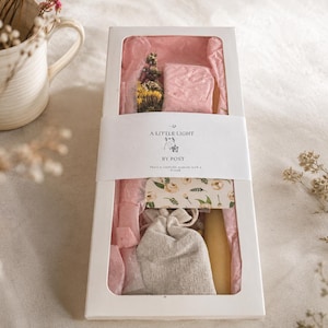 May include: A rectangular white gift box with a clear window containing pink tissue paper, a small bundle of dried flowers, a beige fabric pouch, and a yellow candle. A white paper band features the text A LITTLE LIGHT BY POST.