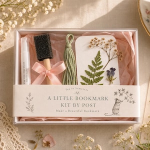 May include: A bookmark craft kit in a clear box containing a small spray bottle, a black foam brush with a pink ribbon, a green cord tassel, and a white card decorated with pressed fern and flower specimens. The box features a paper band labelled A LITTLE BOOKMARK KIT BY POST with a mouse illustration.