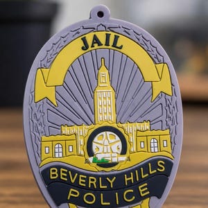 May include: An oval-shaped, light purple silicone badge featuring a yellow banner with the word JAIL at the top. The center displays a yellow building with a tower, and a dark blue banner at the bottom reads BEVERLY HILLS POLICE EST.1906.