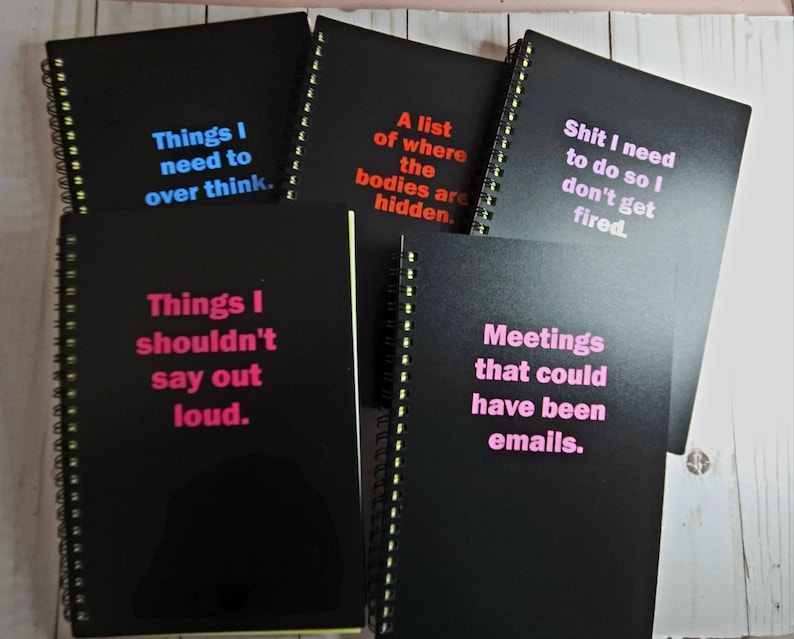 K&ouml;nnte beinhalten: Eine Sammlung schwarzer Spiralnotizb&uuml;cher mit verschiedenen S&auml;tzen in leuchtenden Farben. Die S&auml;tze lauten unter anderem: "Things I need to over think", "A list of where the bodies are hidden" und "Meetings that could have been emails."